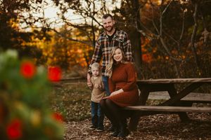 Family portrait on a rustic picnic bench with fall colors and red decorations in background.
