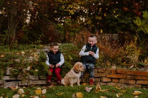 Two young children sit on rustic stone steps with their dog during autumn as fallen leaves scatter the ground.