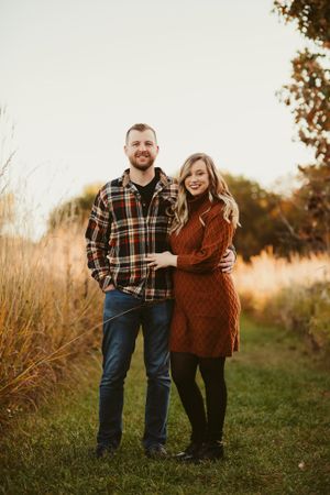 A couple stands together on a grass path during sunset surrounded by autumn foliage.