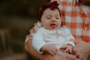 A close-up of a baby in white with a red bow headband showing expression while being held.