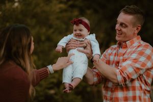 Family playfully lifts baby wearing white outfit and red bow headband during outdoor photo session at sunset.