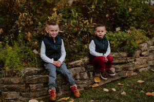 Two children in matching outfits sitting on a stone wall surrounded by fall foliage.