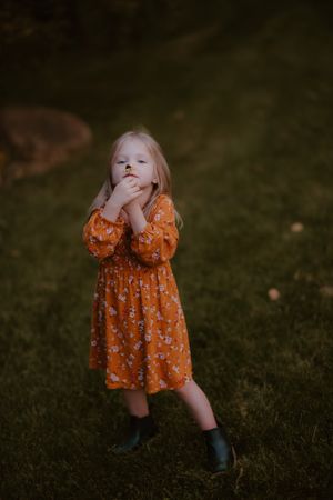 A young child in an orange floral dress and boots strikes a thoughtful pose in the garden.