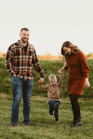 Happy couple swinging child between them while walking in grassy field at sunset.