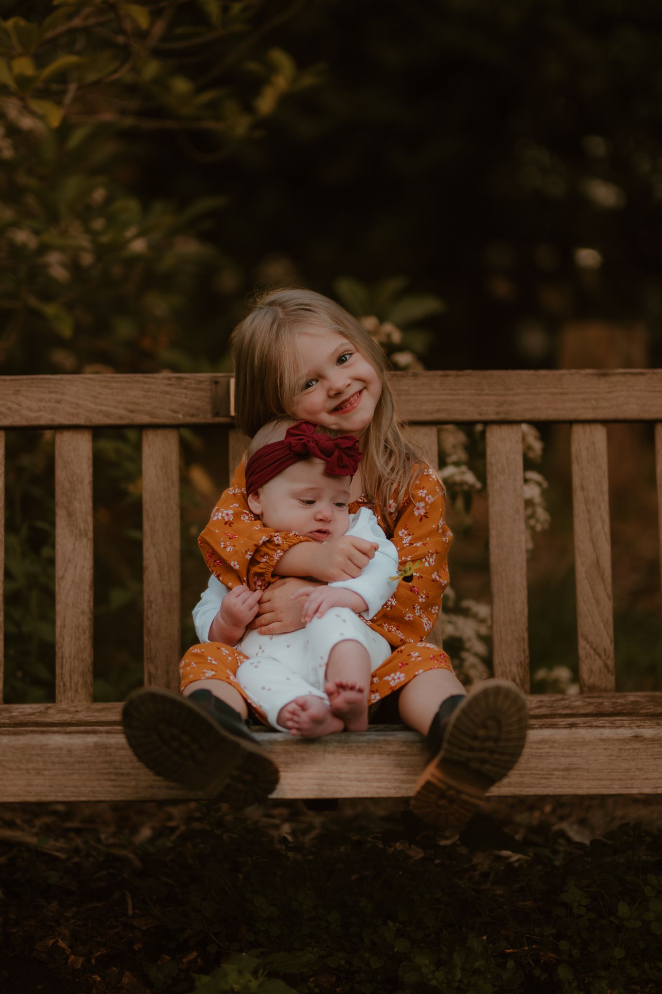 Two children share a sweet moment on a wooden bench, the older wearing orange and younger in white.