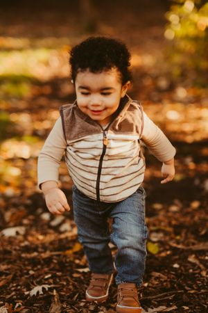 Toddler in striped vest and jeans walking through autumn leaves.