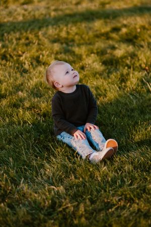 A small child in dark sweater gazes upward while sitting on grass during sunset.