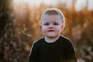A toddler in a dark sweater smiles during a golden hour outdoor photoshoot.