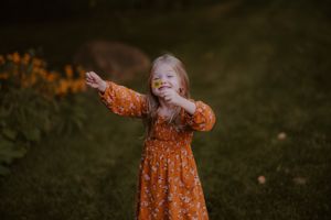 A child in an orange floral dress spreads their arms wide with joy while playing outdoors.