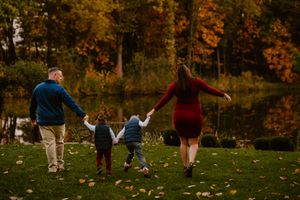 Family holding hands while walking together through a park with autumn trees and a pond in the background.