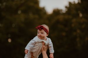 Baby wearing white outfit and red headband being lifted high in the air against blurred nature background.