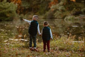 Two children stand by a pond pointing at something in the distance during autumn.