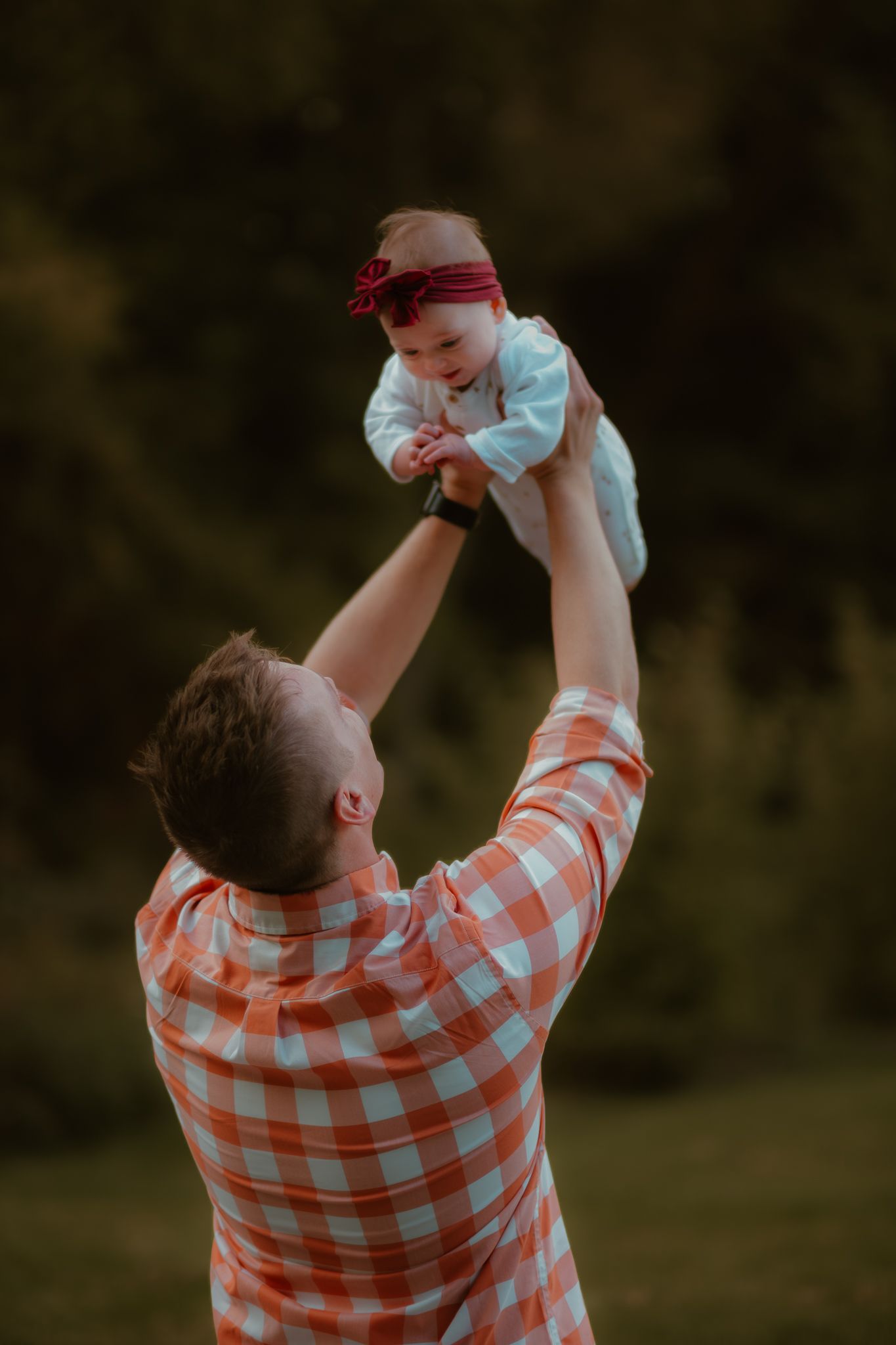 A person in an orange checkered shirt lifts a small baby wearing white and a red bow into the air during an outdoor photoshoot.