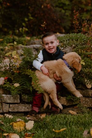 Child in navy vest cuddling with golden retriever puppy on stone steps surrounded by autumn leaves.