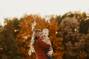 Parent holding child against backdrop of autumn trees at sunset.