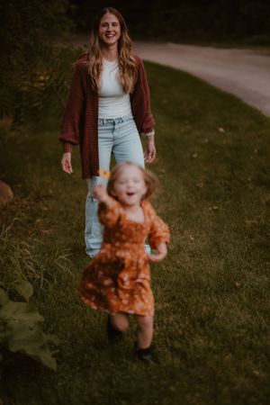 A joyful scene outdoors with a child in orange dress running while someone in jeans and cardigan watches.