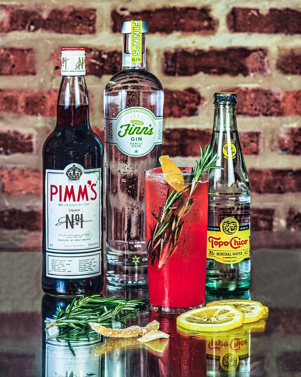 Cocktail ingredients including Pimm's, Hinds gin, and tonic water with citrus garnishes against brick wall.