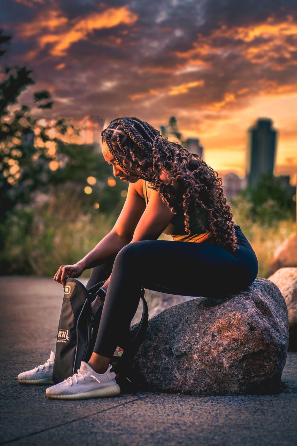Dramatic sunset silhouette photo of someone resting on a rock with curly hair and urban skyline in background.