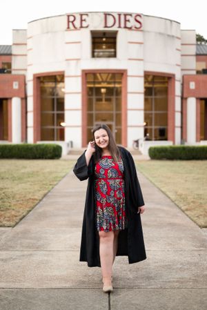 Graduate posing in a black robe and floral dress in front of a collegiate building entrance.