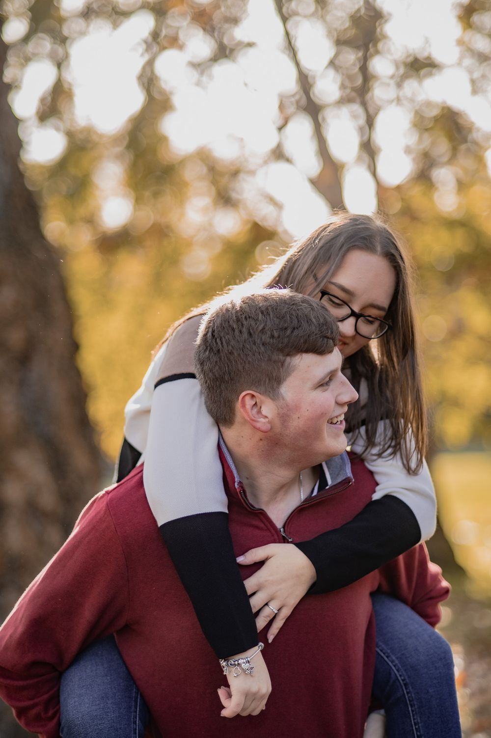 A couple share a playful piggyback ride moment in an autumn park setting with golden bokeh background.