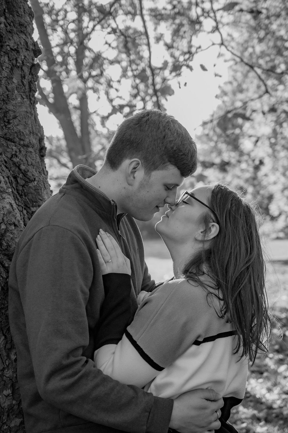 A tender black and white portrait of a couple embracing against a tree in soft natural light.