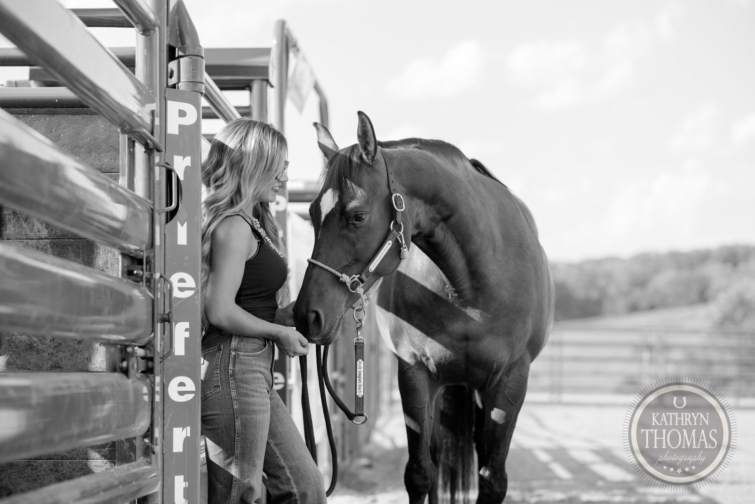 A black and white photo shows someone bonding with their horse near metal stable gates.