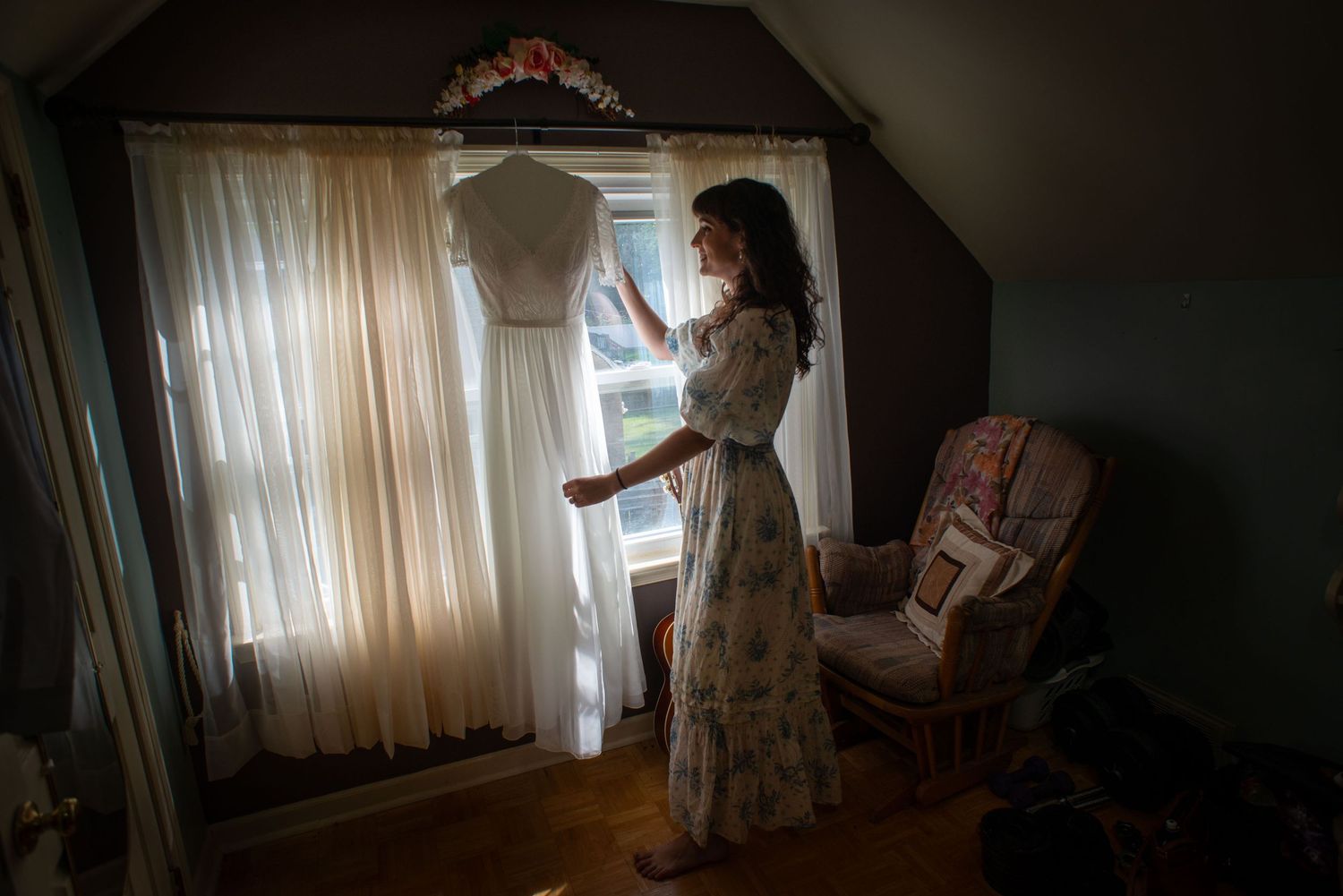 A white bridal dress hangs in a sun-lit room with vintage decor while a figure stands nearby in a floral dress.