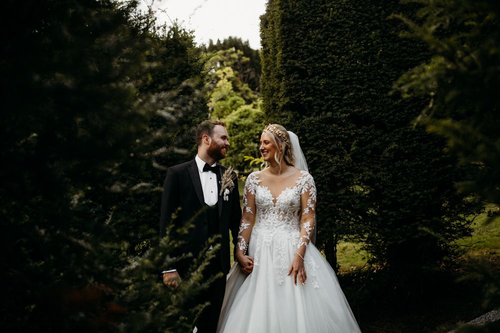Wedding couple standing among green hedges in elegant attire.