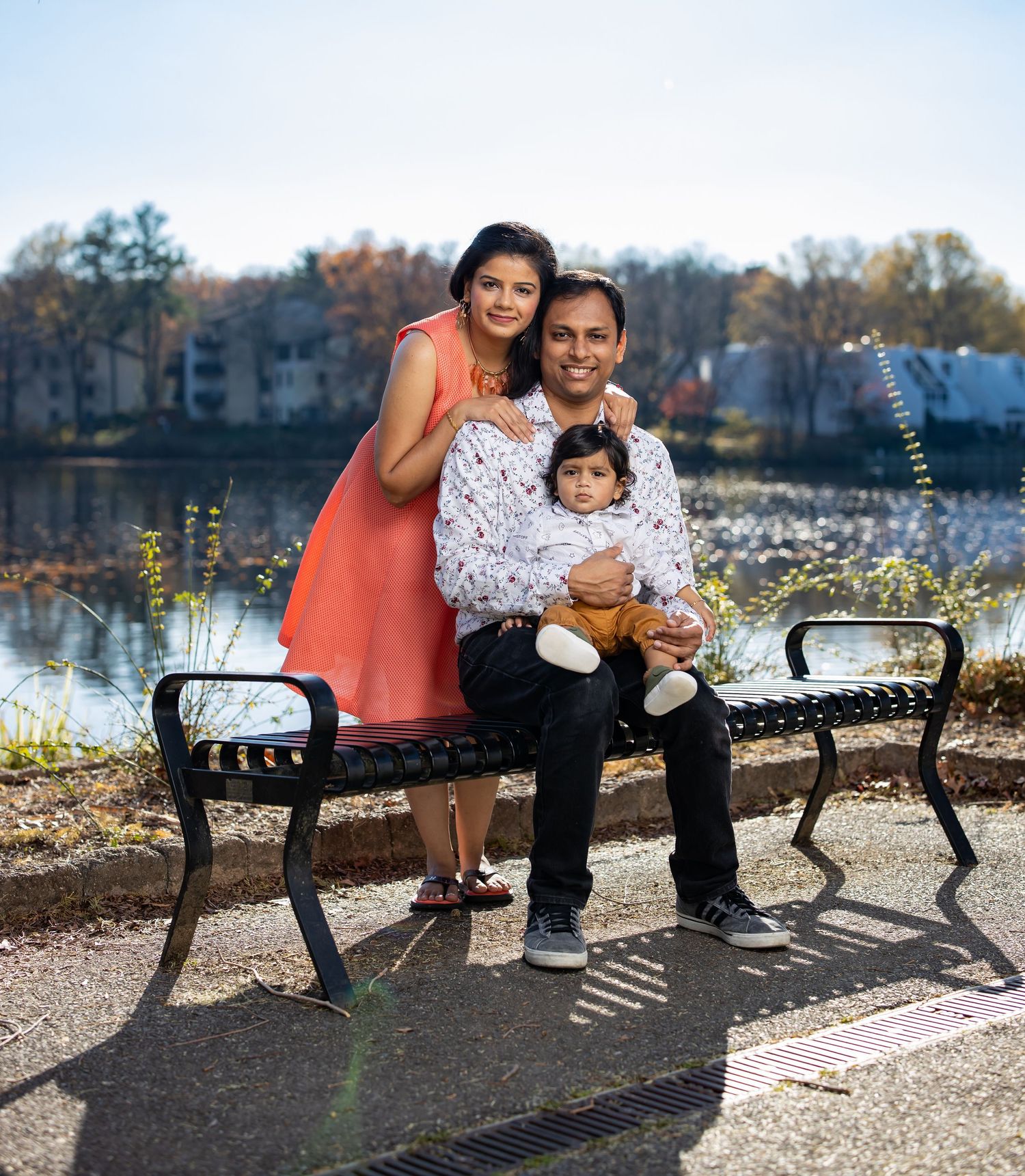 Family sits on park bench near lake during fall season photo shoot.
