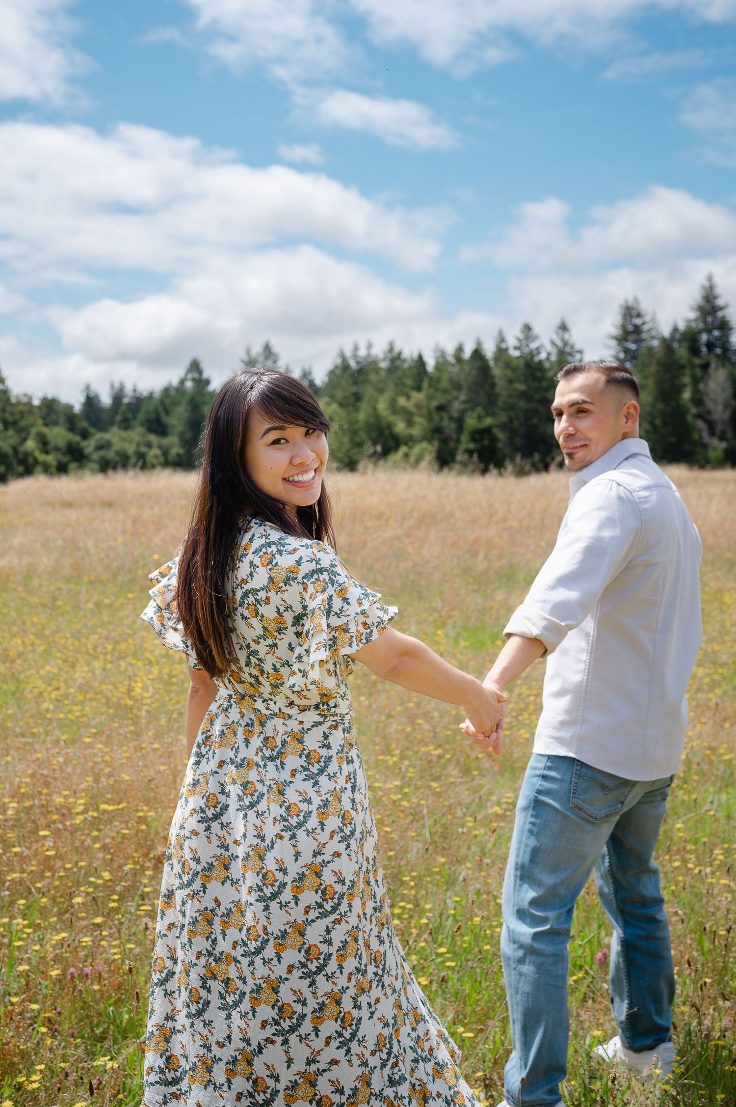 A couple holds hands in a sunlit meadow with forest trees in the background.