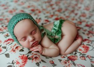 Sleeping newborn baby wearing green knit bonnet posed on floral fabric backdrop.