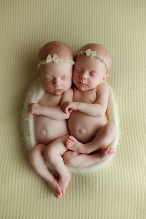 Twin newborn girls in floral headbands cuddling together in a white bowl on green backdrop.