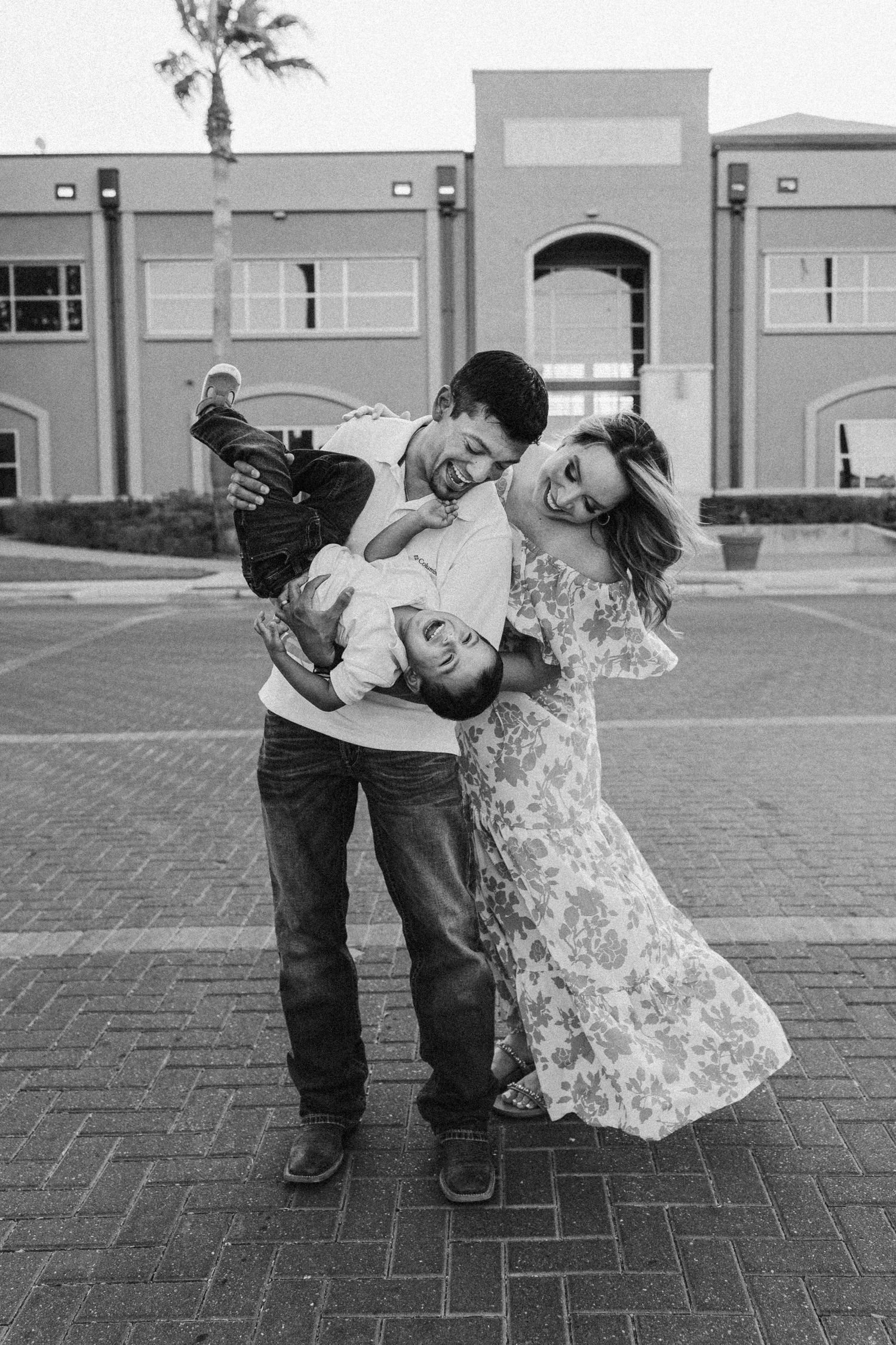 Black and white photo of playful family with two children laughing outdoors on brick plaza.