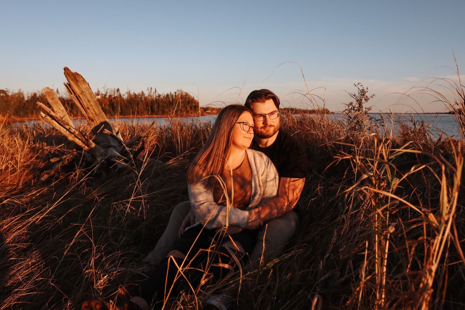 A couple embraces in tall grass during golden hour with water in the background.