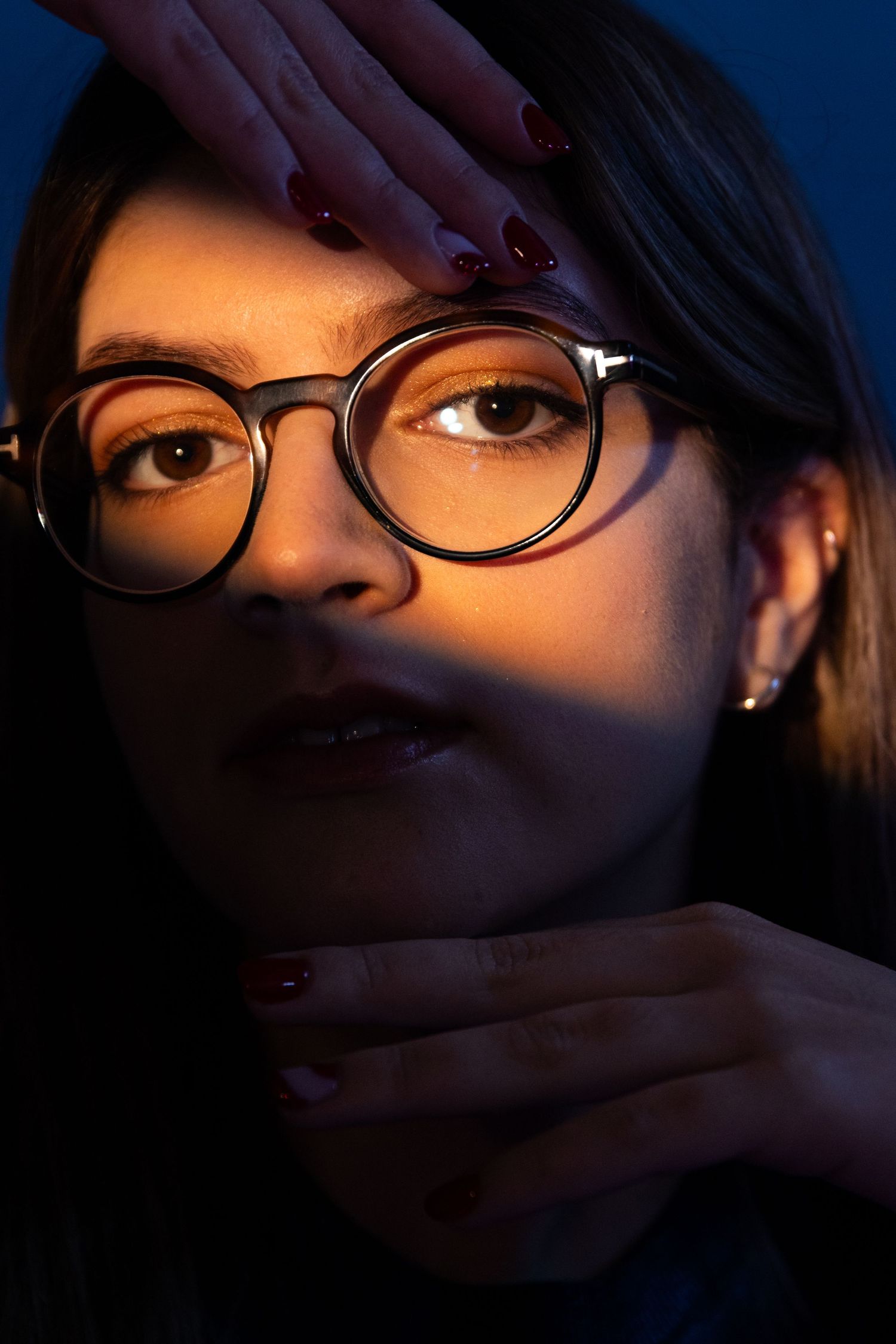 Dramatic close-up portrait with glasses against dark background lit by warm light.