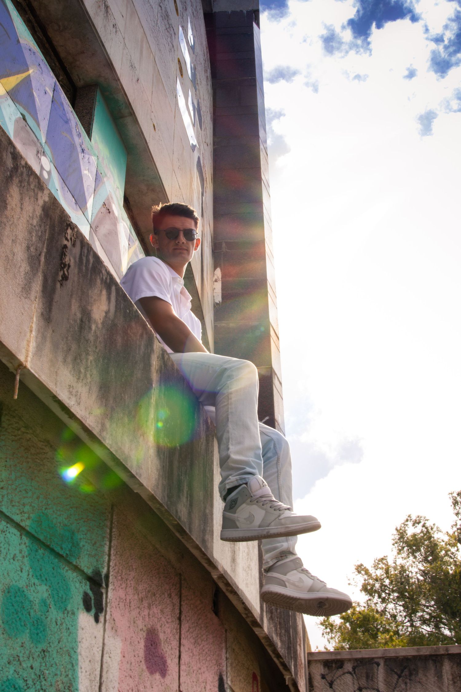 A person in white clothing sits casually on a colorful graffiti wall ledge against a bright sky.