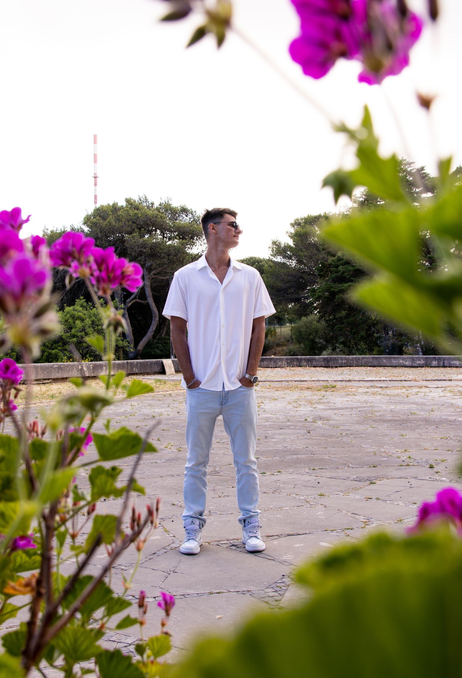 A casual portrait among vibrant pink flowers with greenery and architecture in the background.