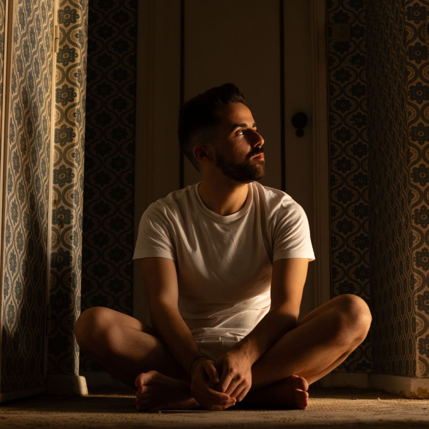 Person in white t-shirt sitting thoughtfully by window in dark room with ornate wall details.