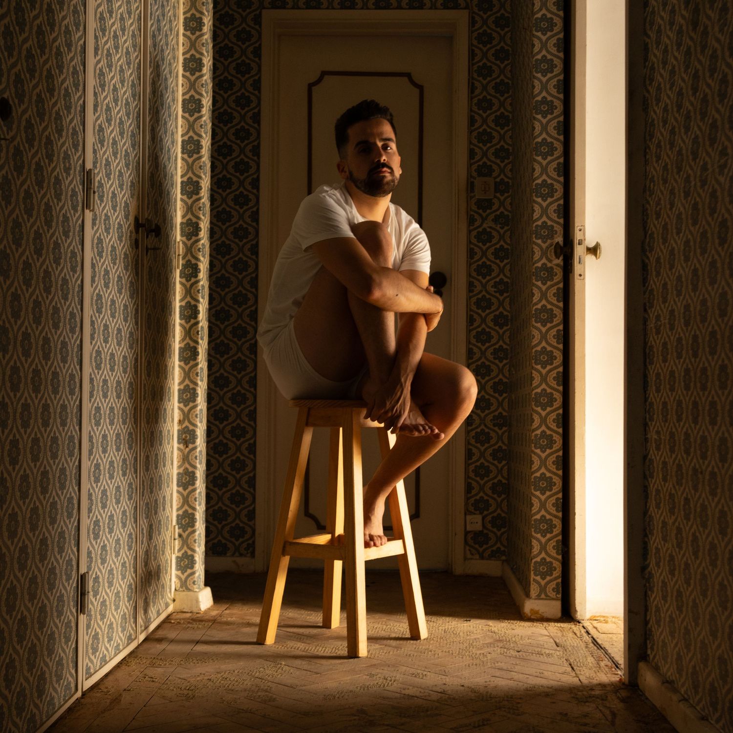 A moody portrait on a wooden stool in a doorway with dramatic lighting and patterned wallpaper.