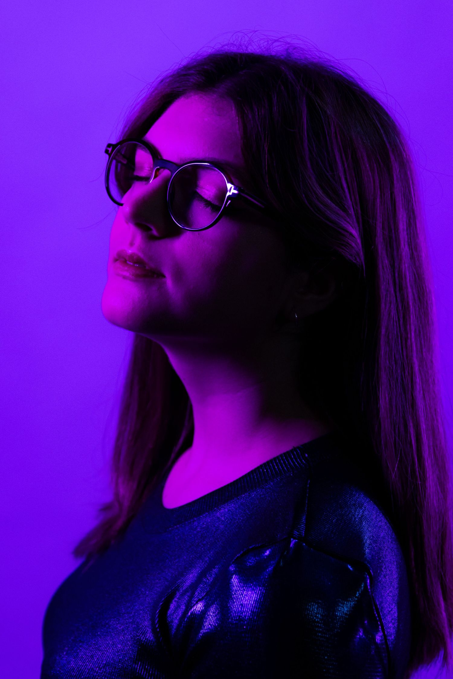 Artistic portrait in purple lighting showing person wearing glasses looking upward.