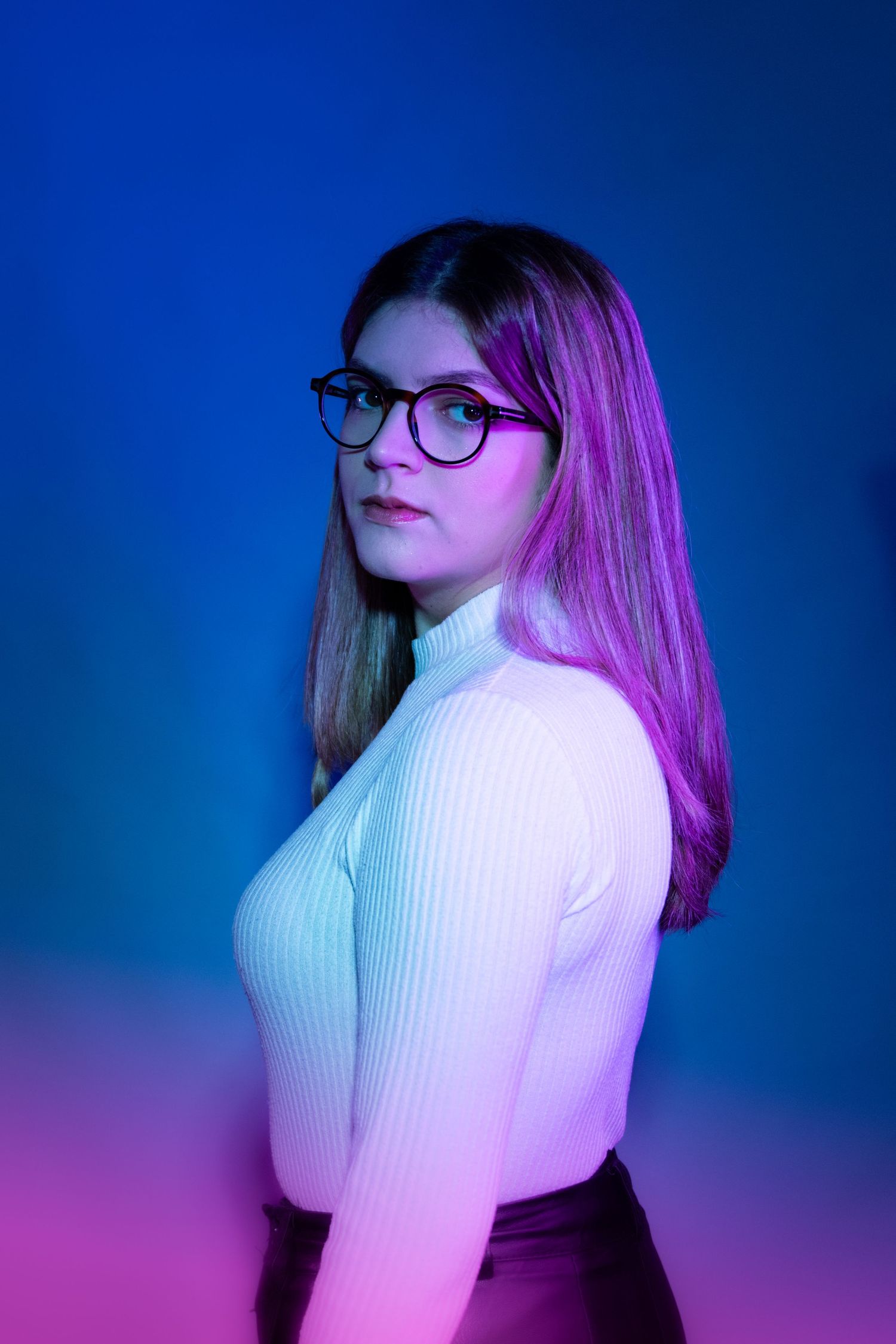Portrait against blue backdrop wearing white turtleneck sweater and glasses with pink lighting.