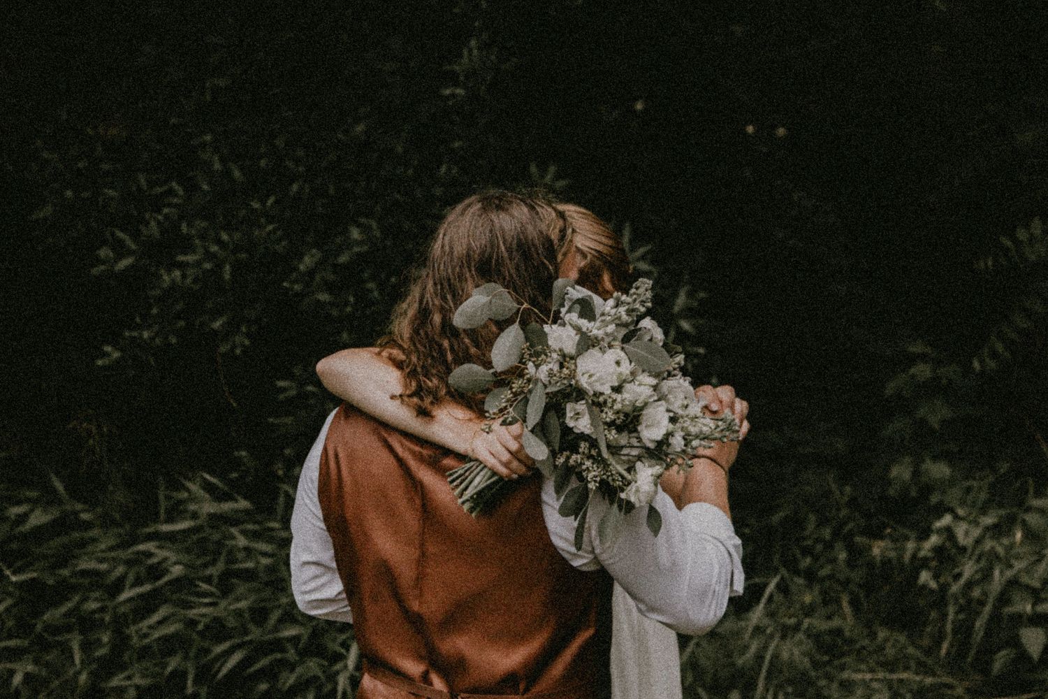 Wedding couple embracing while holding floral bouquet against dark foliage.
