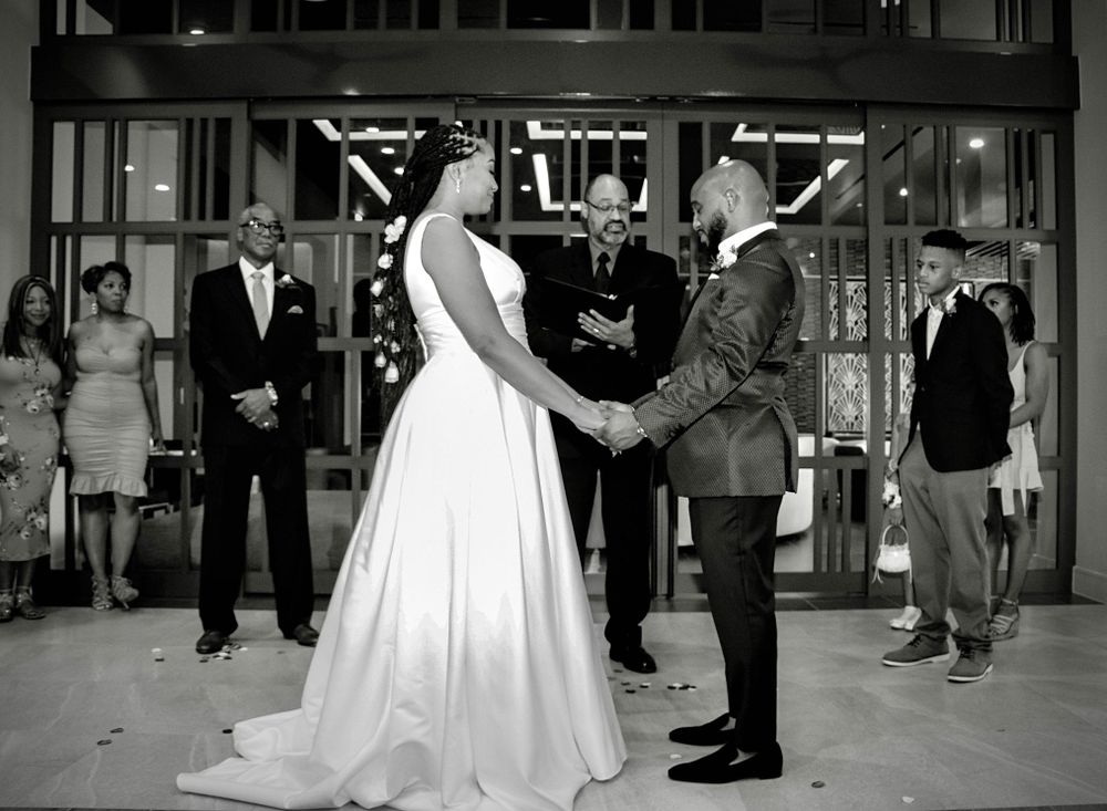 Black and white wedding ceremony photo of couple exchanging vows with guests watching.