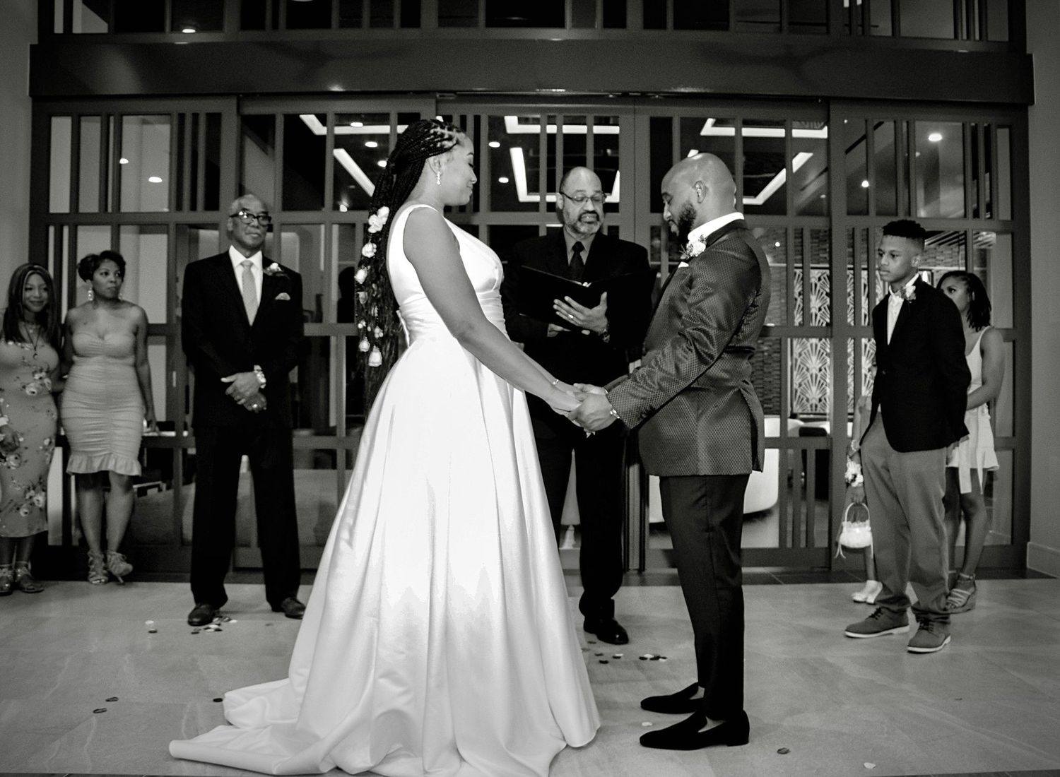 Black and white wedding ceremony photo of couple exchanging vows with guests watching.