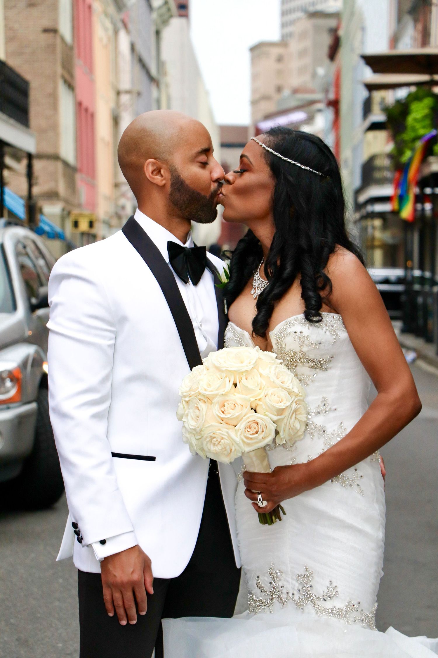 Newlyweds kiss on city street with bride holding white rose bouquet.