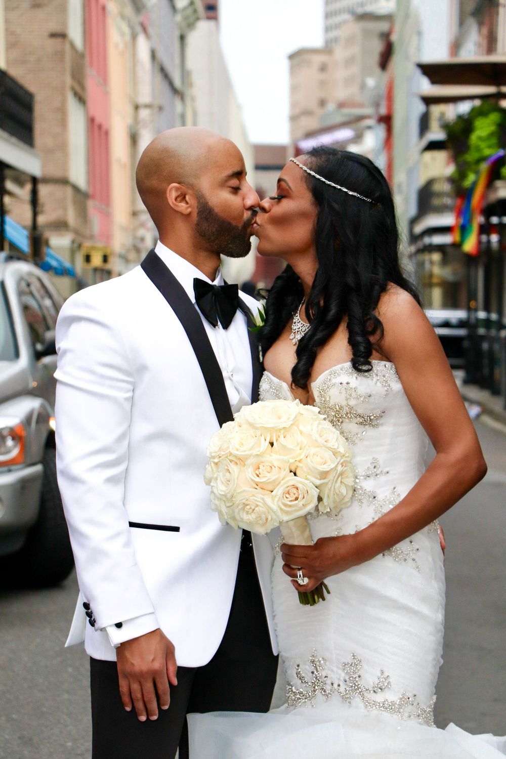 Newlyweds kiss on city street with bride holding white rose bouquet.
