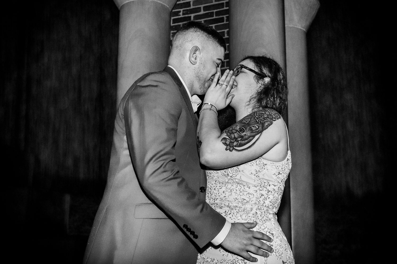 A romantic black and white wedding photo captures a couple kissing under stone columns.