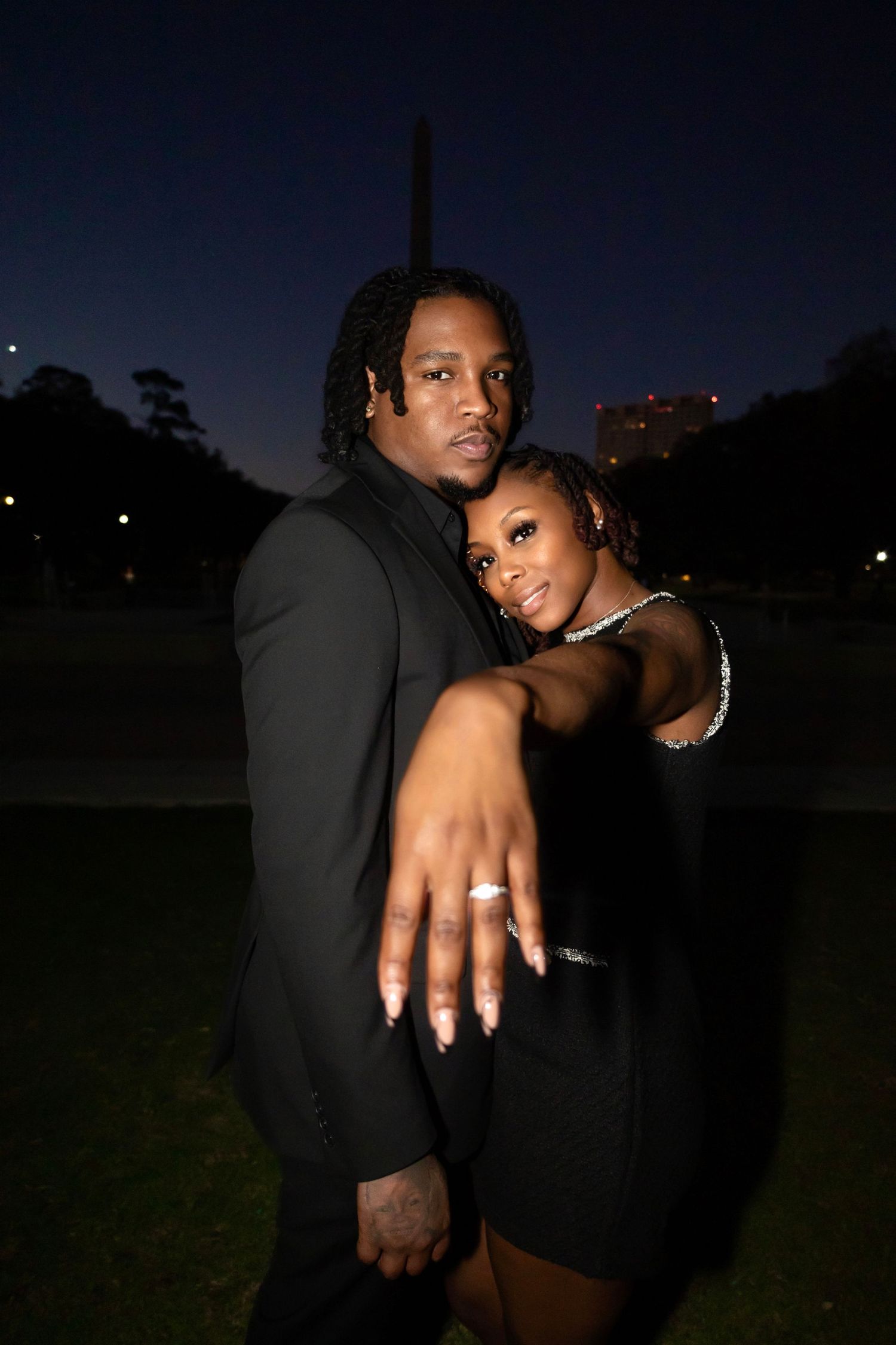 Couple in black outfits showing off engagement rings in nighttime outdoor setting.