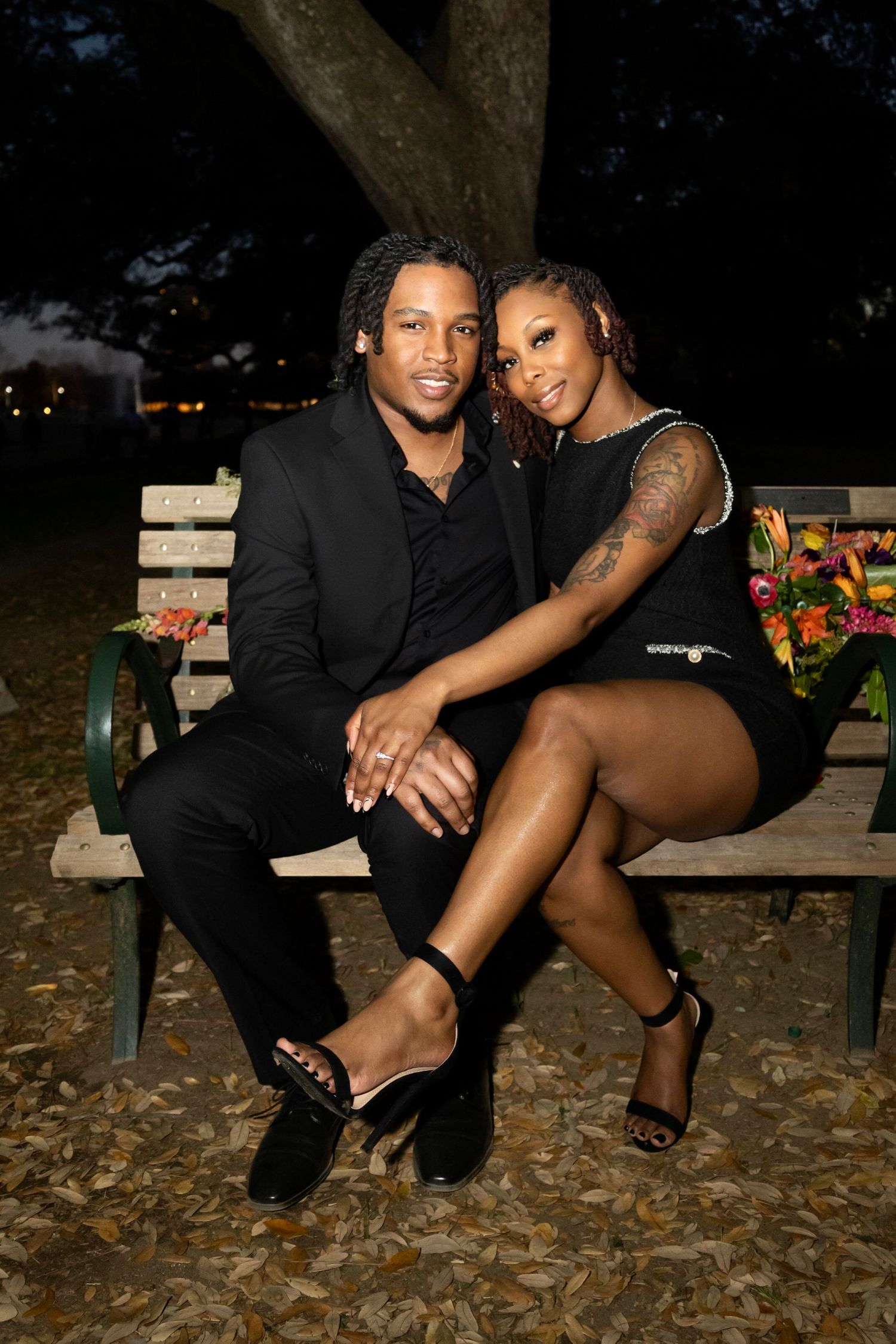 Couple in formal black attire poses romantically on park bench at dusk.
