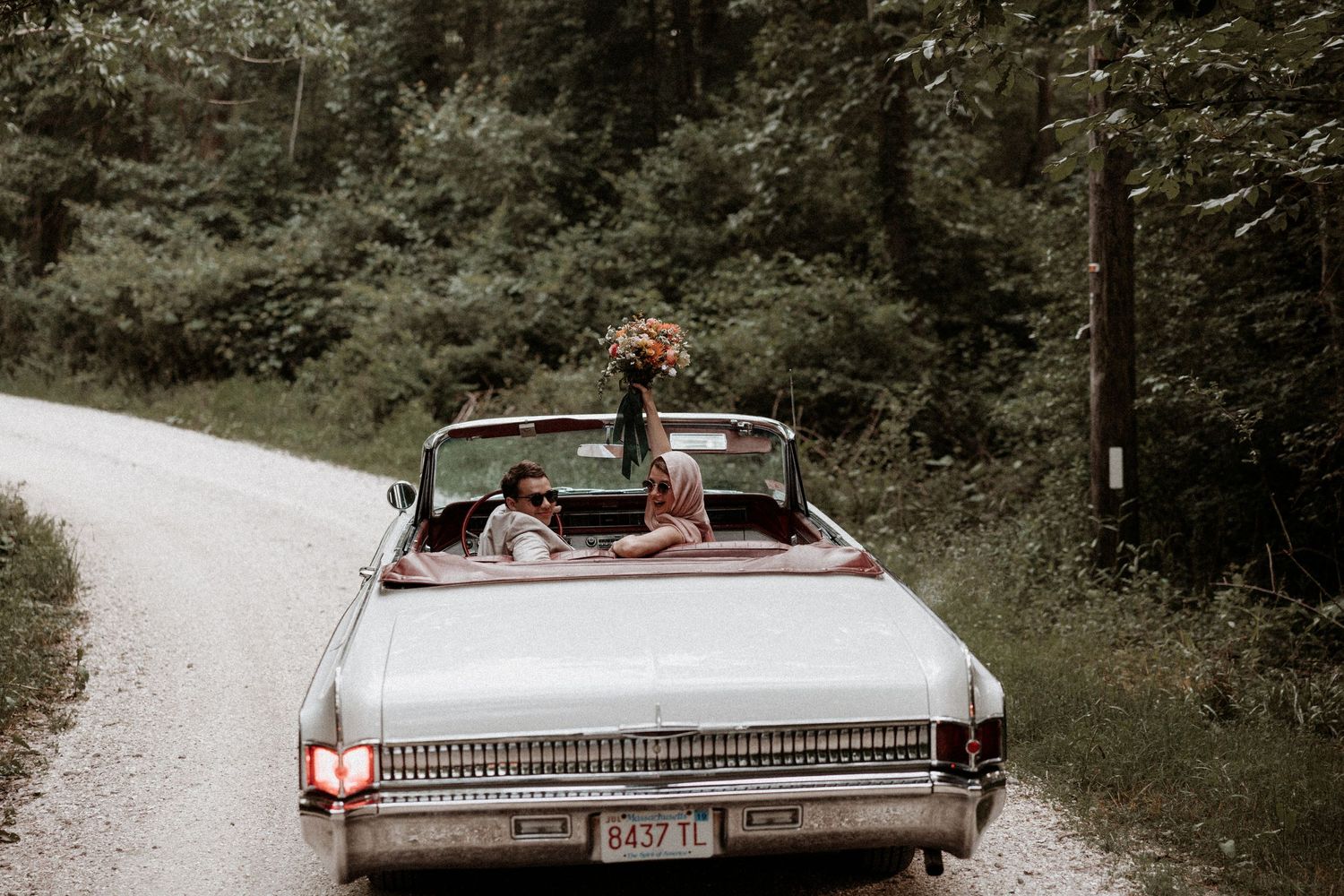 Couple rides in vintage white convertible down wooded gravel road with floral bouquet on trunk. Westport Ma. Miranda Photo & Co